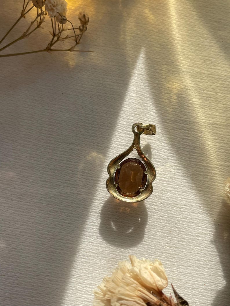 May include: A gold-toned pendant featuring an oval-shaped, translucent amber-colored gemstone set within a curved, open-work metal frame. The pendant includes a small circular bail at the top for attachment to a chain or cord.