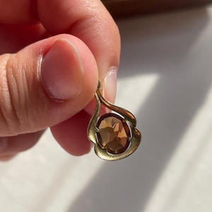 May include: A gold-toned metal pendant featuring a teardrop-shaped frame with a smooth, curved design. The center of the pendant holds a single, oval-cut, faceted brown gemstone that reflects light, creating a warm, amber-colored appearance.