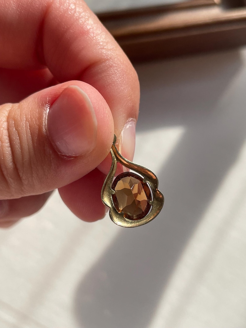 May include: A gold-toned metal pendant featuring a teardrop-shaped frame with a smooth, curved design. The center of the pendant holds a single, oval-cut, faceted brown gemstone that reflects light, creating a warm, amber-colored appearance.
