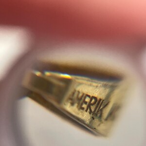 May include: A gold-toned metal jewelry component featuring a stamped inscription that reads AMERIK. The object has a triangular, tapered shape with a smooth, reflective surface and a small loop at the top for attachment to a chain or cord.