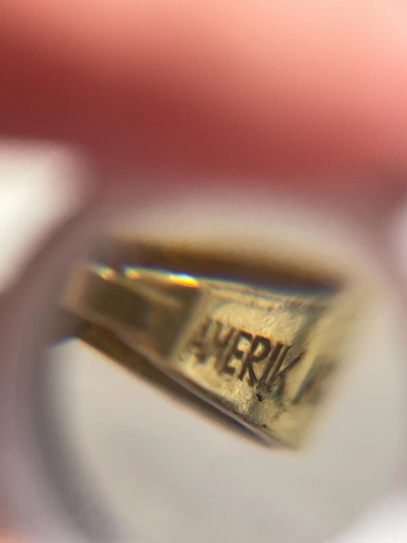 May include: A gold-toned metal jewelry component featuring a stamped inscription that reads AMERIK. The object has a triangular, tapered shape with a smooth, reflective surface and a small loop at the top for attachment to a chain or cord.