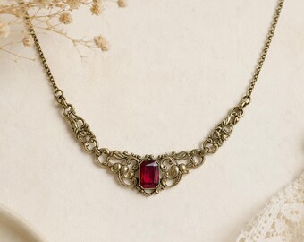 May include: A necklace featuring a central rectangular deep red gemstone set in an ornate antique brass-toned metal frame with intricate scrollwork patterns, connected to a delicate matching metal chain on both sides of the decorative centerpiece.