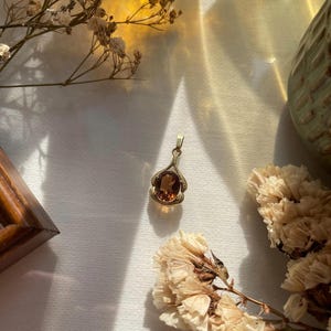 May include: A gold pendant featuring an oval-shaped, deep amber-colored gemstone set within a decorative metal frame. The pendant has a small bail at the top for attachment to a chain, showcasing a polished finish and a warm, translucent appearance.