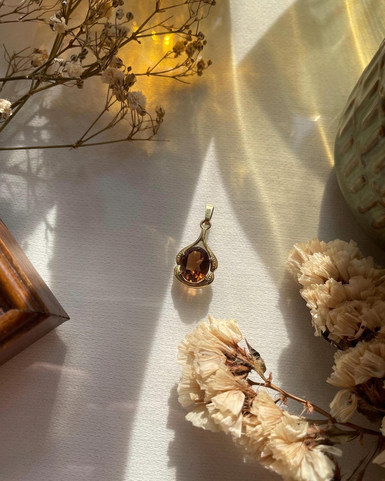 May include: A gold pendant featuring an oval-shaped, deep amber-colored gemstone set within a decorative metal frame. The pendant has a small bail at the top for attachment to a chain, showcasing a polished finish and a warm, translucent appearance.