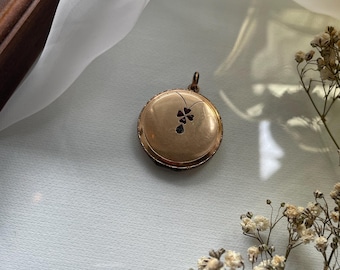 May include: A round gold-toned locket pendant featuring a small, dark four-leaf clover inlay at the center. The locket has a smooth, polished surface with a delicate metal loop at the top for attachment to a chain or cord, showcasing a classic design.