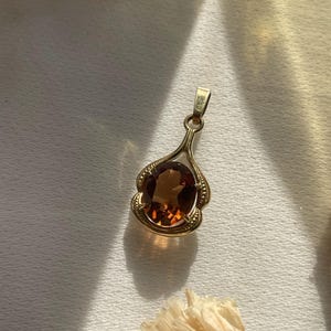 May include: A gold pendant featuring an oval-shaped, deep amber-colored gemstone set within a decorative, scalloped gold frame. The pendant includes a small, integrated gold bail at the top for attachment to a chain, showcasing a polished finish.