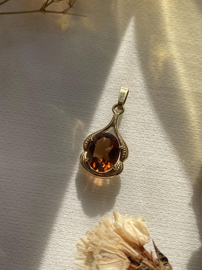 May include: A gold pendant featuring an oval-shaped, deep amber-colored gemstone set within a decorative, scalloped gold frame. The pendant includes a small, integrated gold bail at the top for attachment to a chain, showcasing a polished finish.