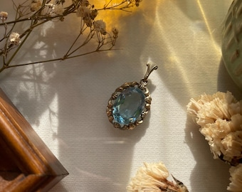 May include: An oval-shaped pendant featuring a faceted light blue gemstone set in an ornate, antique-toned metal frame with a decorative bail at the top. The metalwork displays a textured, scalloped edge surrounding the central blue stone.