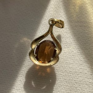 May include: A gold-toned pendant featuring an oval-shaped, faceted brown gemstone set within a curved, open-frame metal setting. The pendant includes a small, integrated bail at the top for attachment to a chain, showcasing a polished metallic finish.
