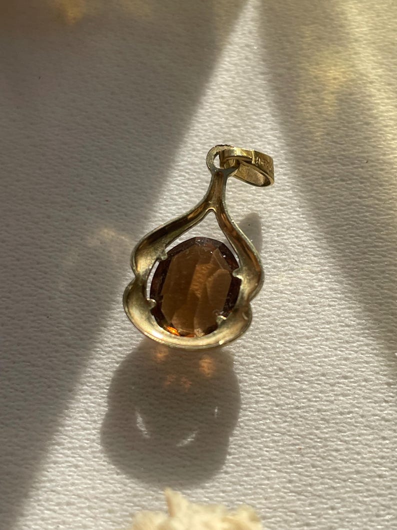 May include: A gold-toned pendant featuring an oval-shaped, faceted brown gemstone set within a curved, open-frame metal setting. The pendant includes a small, integrated bail at the top for attachment to a chain, showcasing a polished metallic finish.