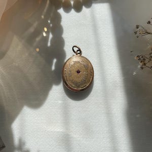 Könnte beinhalten: Ein ovaler Medaillon-Anhänger aus goldfarbenem Metall mit einem aufwendig gravierten Randmuster und einem kleinen, mittig platzierten violetten Edelstein. Das Medaillon besitzt eine runde Öse an der Oberseite zur Befestigung an einer Kette.