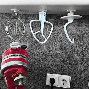 Könnte beinhalten: Ein roter KitchenAid-Küchenmixer mit drei Aufsätzen, die darüber hängen. Zu den Aufsätzen gehören ein Schneebesen, ein Flachrührer und ein Knethaken. Der Mixer und die Aufsätze befinden sich vor einer grau strukturierten Wand.