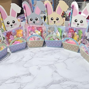 May include: Easter gift baskets featuring bunny cutouts, filled with sweets and toys. Each basket has a small handle and is filled with colourful candies and small toys. The bunny cutouts have pink ears and cute faces.