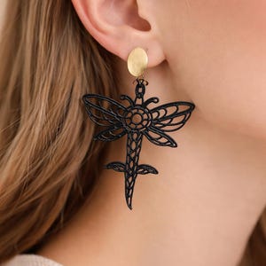 Può includere: Orecchini a forma di libellula neri con borchia color oro. Gli orecchini presentano un design traforato e dettagliato. Le ali e il corpo della libellula sono finemente lavorati, creando un accessorio delicato ed elegante. Questi orecchini sono un'aggiunta unica.