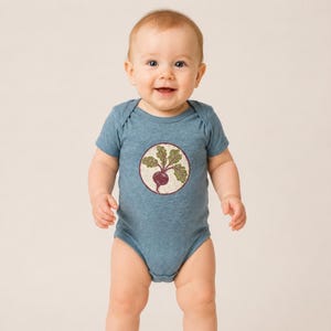 May include: A heathered blue short-sleeve baby bodysuit featuring a circular graphic on the chest. The graphic depicts a purple beet with green leaves inside a cream-coloured circle with a dark red border, centred on the front of the garment.
