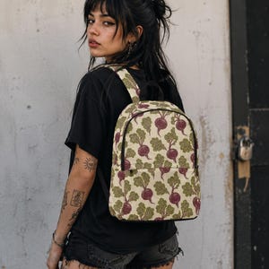 May include: A backpack featuring a repeating pattern of purple beets with green leafy tops on a cream-colored background. The bag has a main zippered compartment, a front zippered pocket, and adjustable shoulder straps with black trim.