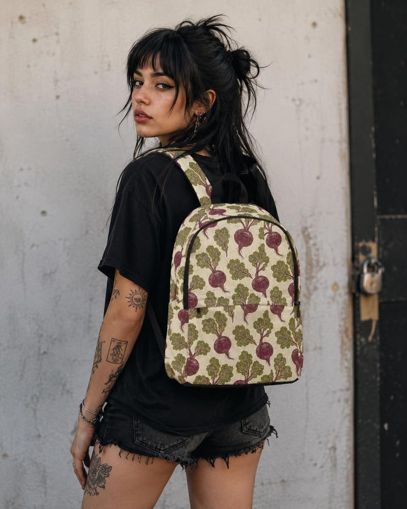 May include: A backpack featuring a repeating pattern of purple beets with green leafy tops on a cream-colored background. The bag has a main zippered compartment, a front zippered pocket, and adjustable shoulder straps with black trim.