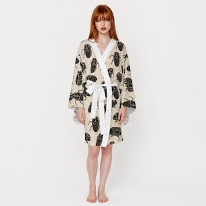 May include: A beige kimono-style robe featuring a repeating black beetle print. The garment has long, wide sleeves, a solid white trim along the front opening, and a matching white fabric tie belt secured at the waist to close the front.