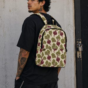 May include: A backpack featuring a repeating pattern of purple beetroots with green leafy tops on a cream-colored background. The bag has black trim, a top handle, and adjustable shoulder straps, with a zippered front pocket and a main compartment.