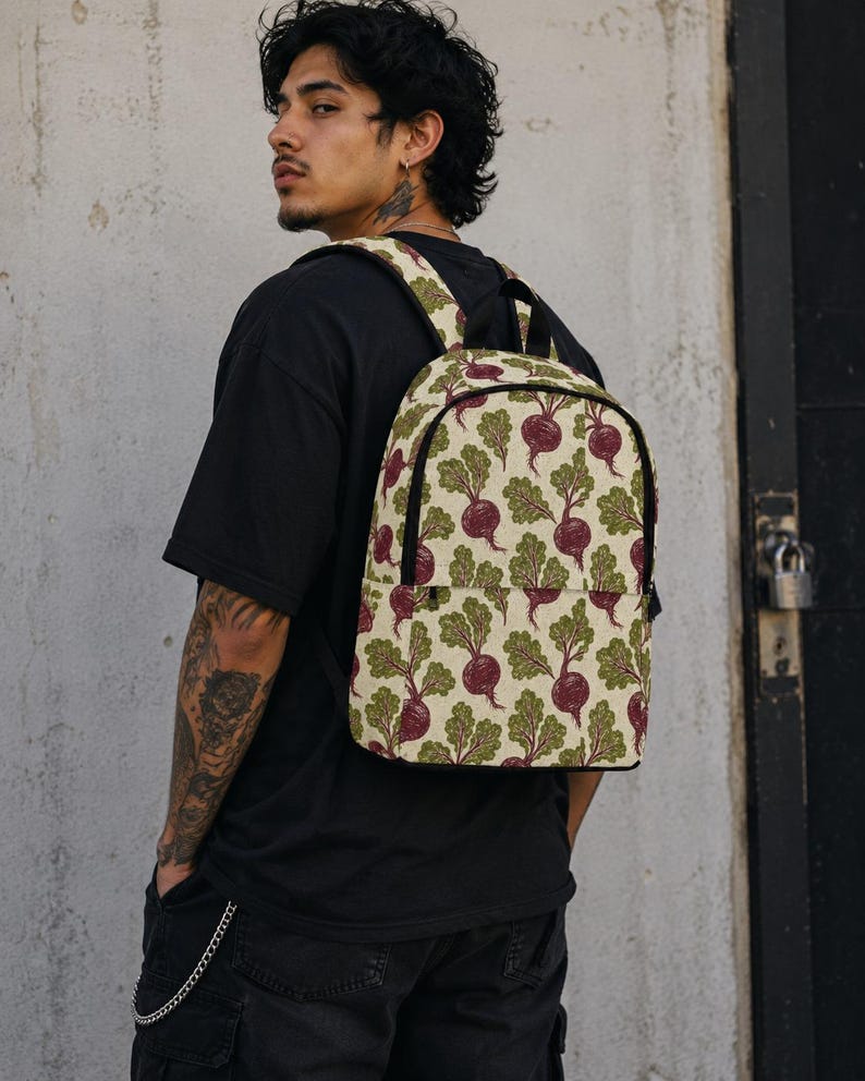 May include: A backpack featuring a repeating pattern of purple beetroots with green leafy tops on a cream-colored background. The bag has black trim, a top handle, and adjustable shoulder straps, with a zippered front pocket and a main compartment.