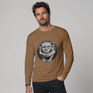 May include: A long-sleeve crew neck t-shirt in a solid tan color. The center of the chest features a circular graphic print containing a detailed black and white illustration of a seated, textured creature with large ears and a stoic facial expression.