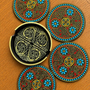 May include: A set of three circular coasters featuring intricate black, red, and yellow Celtic knotwork patterns with bright blue accents, accompanied by a black circular holder with a gold-colored Celtic knot design on its interior base.