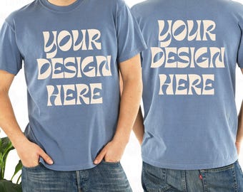Puede incluir: Una camiseta de manga corta en color azul polvoriento con el texto "YOUR DESIGN HERE" impreso en una fuente grande, ondulada y de estilo retro de color crema tanto en la parte delantera como en la trasera. Tiene cuello redondo y corte estándar.