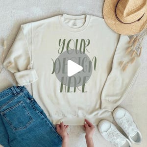 Puede incluir: Una sudadera de cuello redondo color crema con el texto "YOUR DESIGN HERE" en letras verde oliva, combinada con vaqueros azules, zapatillas blancas y un sombrero de paja. La sudadera tiene mangas largas y cuello, puños y dobladillo acanalados.
