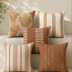 May include: A set of five square throw pillows in shades of terracotta, cream, and tan. The designs include geometric fan patterns, vertical stripes, a central column of horizontal dashes, and delicate dotted leaf-like vertical line motifs.