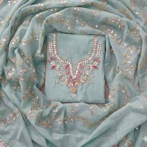 May include: A light teal salwar kameez set. The top features intricate embroidery around the neckline with pink and gold accents. The matching dupatta and top are adorned with gold leaf-like embellishments, creating an elegant and festive look.