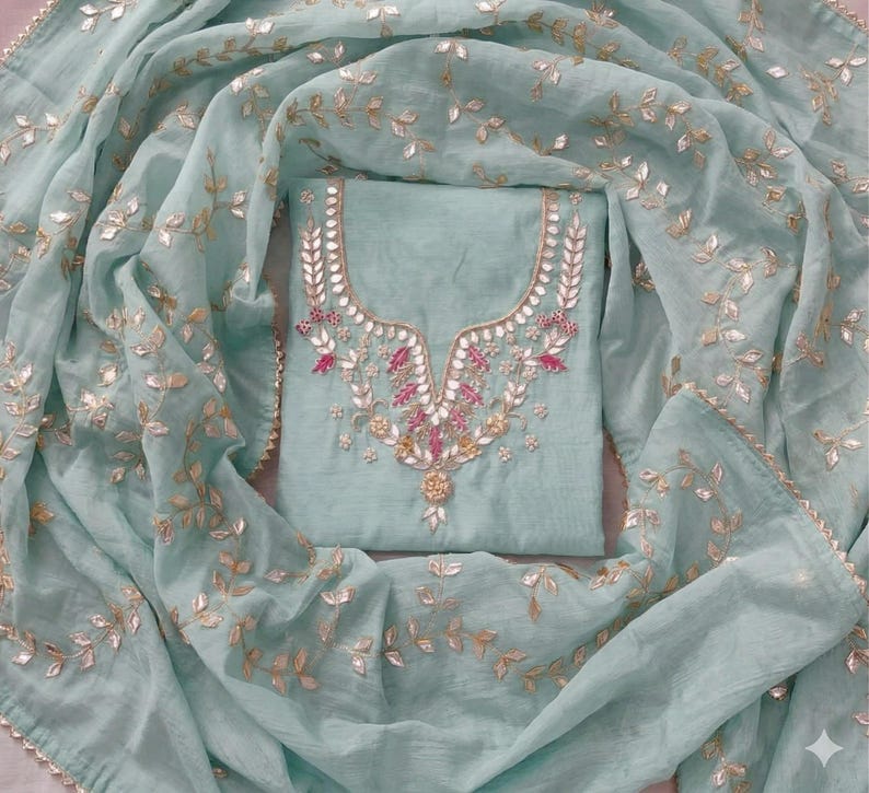 May include: A light teal salwar kameez set. The top features intricate embroidery around the neckline with pink and gold accents. The matching dupatta and top are adorned with gold leaf-like embellishments, creating an elegant and festive look.