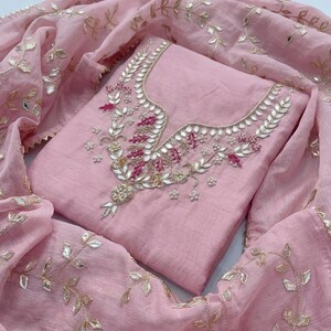 May include: A light pink salwar kameez ensemble. The top features intricate embroidery with silver and pink accents around the neckline. A matching dupatta with gold leaf-like embellishments drapes around the outfit, creating a cohesive look.