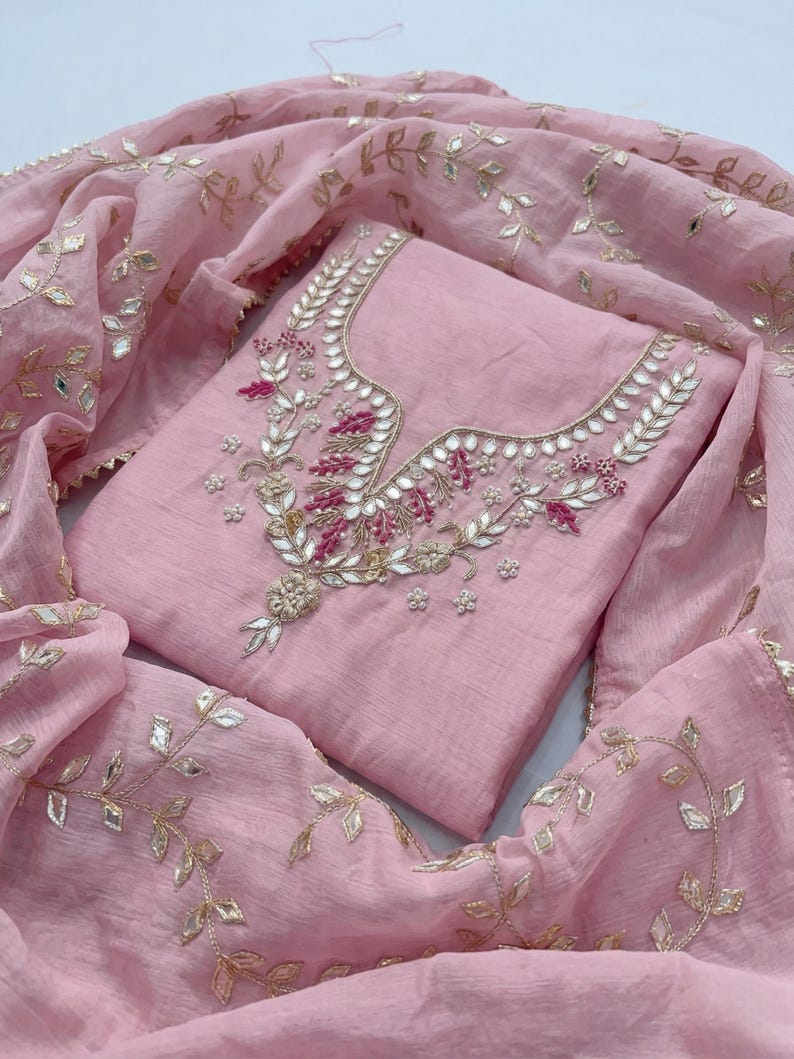 May include: A light pink salwar kameez ensemble. The top features intricate embroidery with silver and pink accents around the neckline. A matching dupatta with gold leaf-like embellishments drapes around the outfit, creating a cohesive look.