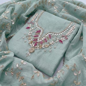 May include: A light teal-colored embroidered salwar kameez set. The top features intricate embroidery with silver and pink accents around the neckline. The matching dupatta is adorned with similar leaf-shaped embroidery.