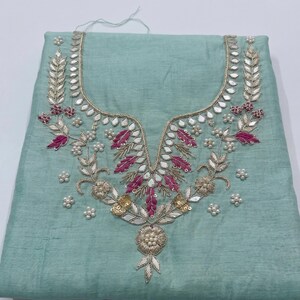 May include: A light teal fabric with an ornate embroidered neckline. The embroidery features silver, gold, and pink thread, along with small white pearl-like beads. The design is symmetrical and floral, with a detailed, elegant appearance.