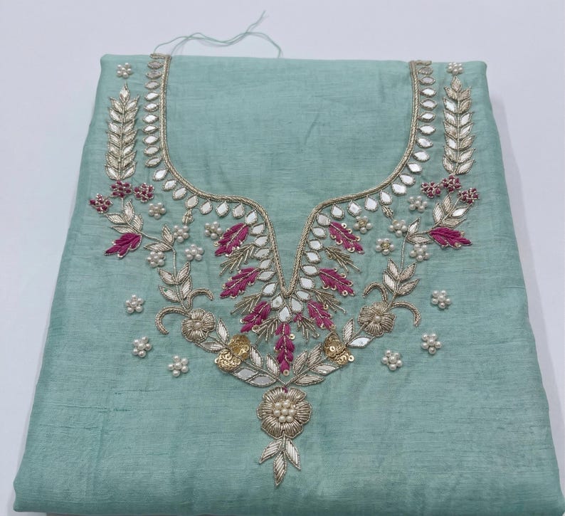 May include: A light teal fabric with an ornate embroidered neckline. The embroidery features silver, gold, and pink thread, along with small white pearl-like beads. The design is symmetrical and floral, with a detailed, elegant appearance.
