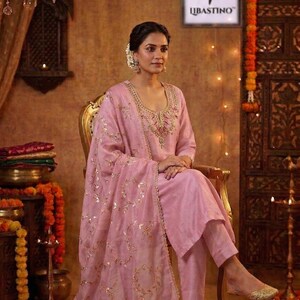 May include: A light pink salwar kameez set with gold embroidery. The set includes a long-sleeved tunic, matching pants, and a dupatta. The outfit is worn with gold-colored flats. The background features traditional Indian decor.