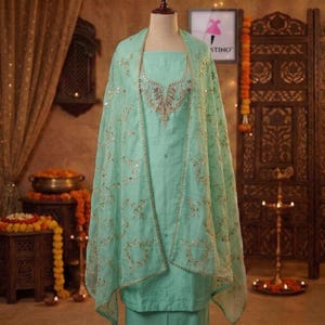 May include: A light green salwar kameez set with intricate gold embroidery and embellishments. The set includes a tunic, pants, and a sheer dupatta. The outfit is displayed on a mannequin, showcasing the traditional Indian design.