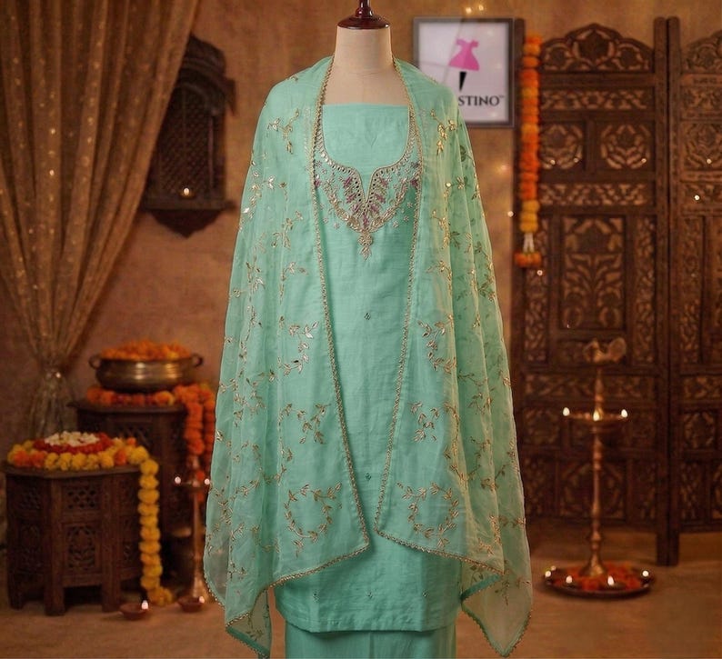 May include: A light green salwar kameez set with intricate gold embroidery and embellishments. The set includes a tunic, pants, and a sheer dupatta. The outfit is displayed on a mannequin, showcasing the traditional Indian design.