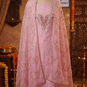 May include: A light pink salwar kameez ensemble on a mannequin. The outfit includes a tunic, pants, and a sheer dupatta with gold leaf-like embroidery. The tunic features intricate embroidery around the neckline. The outfit is displayed in a well-lit setting.