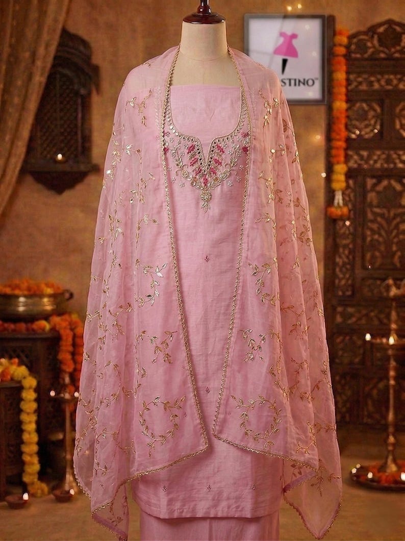 May include: A light pink salwar kameez ensemble on a mannequin. The outfit includes a tunic, pants, and a sheer dupatta with gold leaf-like embroidery. The tunic features intricate embroidery around the neckline. The outfit is displayed in a well-lit setting.