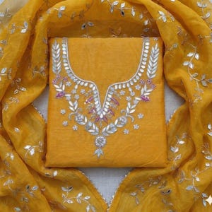 May include: Mustard yellow salwar kameez set with intricate silver and purple embroidery on the neckline. The matching dupatta features silver leaf accents. The fabric has a slight sheen, creating a luxurious look.