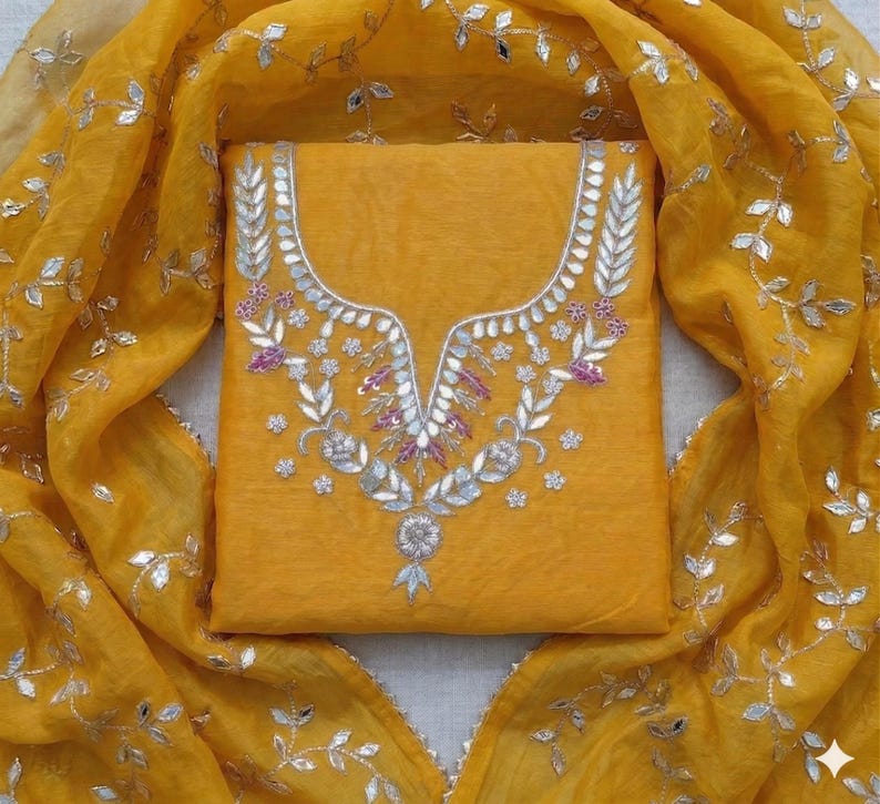 May include: Mustard yellow salwar kameez set with intricate silver and purple embroidery on the neckline. The matching dupatta features silver leaf accents. The fabric has a slight sheen, creating a luxurious look.