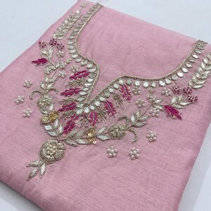 May include: A light pink fabric with intricate embroidery. The embroidery features silver and gold metallic thread, small mirrors, and accents of fuchsia and white beads. The design is concentrated around the neckline.