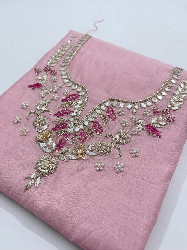 May include: A light pink fabric with intricate embroidery. The embroidery features silver and gold metallic thread, small mirrors, and accents of fuchsia and white beads. The design is concentrated around the neckline.