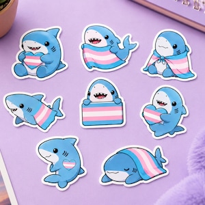 May include: A collection of eight die-cut stickers featuring cartoon blue sharks in various poses, each incorporating the light blue, pink, and white colors of the transgender pride flag in the form of hearts, capes, or rectangular banners.