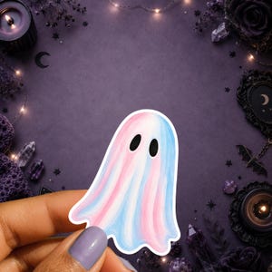 May include: A die-cut sticker featuring a ghost illustration with a watercolour-style blend of pastel pink and light blue vertical stripes. The ghost has two simple black oval eyes and a white border outline, presented on a flat surface.