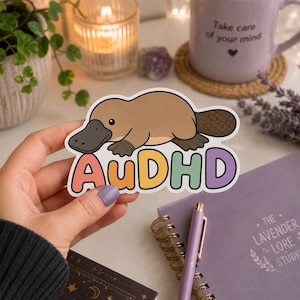 Op de afbeelding: Een gestanste sticker met een cartoon vogelbekdier in bruintinten met een donkere snavel en staart. Onder het vogelbekdier staat de tekst AuDHD in grote, afgeronde, kleurrijke letters in koraal, oranje, geel, groen en paars gedrukt.