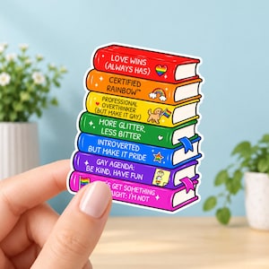 Pode incluir: Um adesivo com uma pilha de sete livros coloridos em ordem de arco-íris. As lombadas exibem textos incluindo LOVE WINS, CERTIFIED RAINBOW, PROFESSIONAL OVERTHINKER, MORE GLITTER, LESS BITTER, INTROVERTED BUT MAKE IT PRIDE, GAY AGENDA e LET'S GET SOMETHING STRAIGHT: I'M NOT.