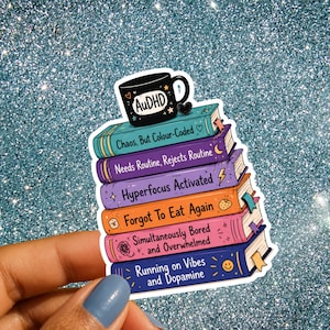 Op de afbeelding: Een sticker met een stapel van zes kleurrijke boeken bekroond met een zwarte mok met het label AuDHD. De boekruggen bevatten de teksten: Chaos, But Colour-Coded, Needs Routine, Rejects Routine, Hyperfocus Activated, Forgot To Eat Again, Simultaneously Bored and Overwhelmed en Running on Vibes and Dopamine.